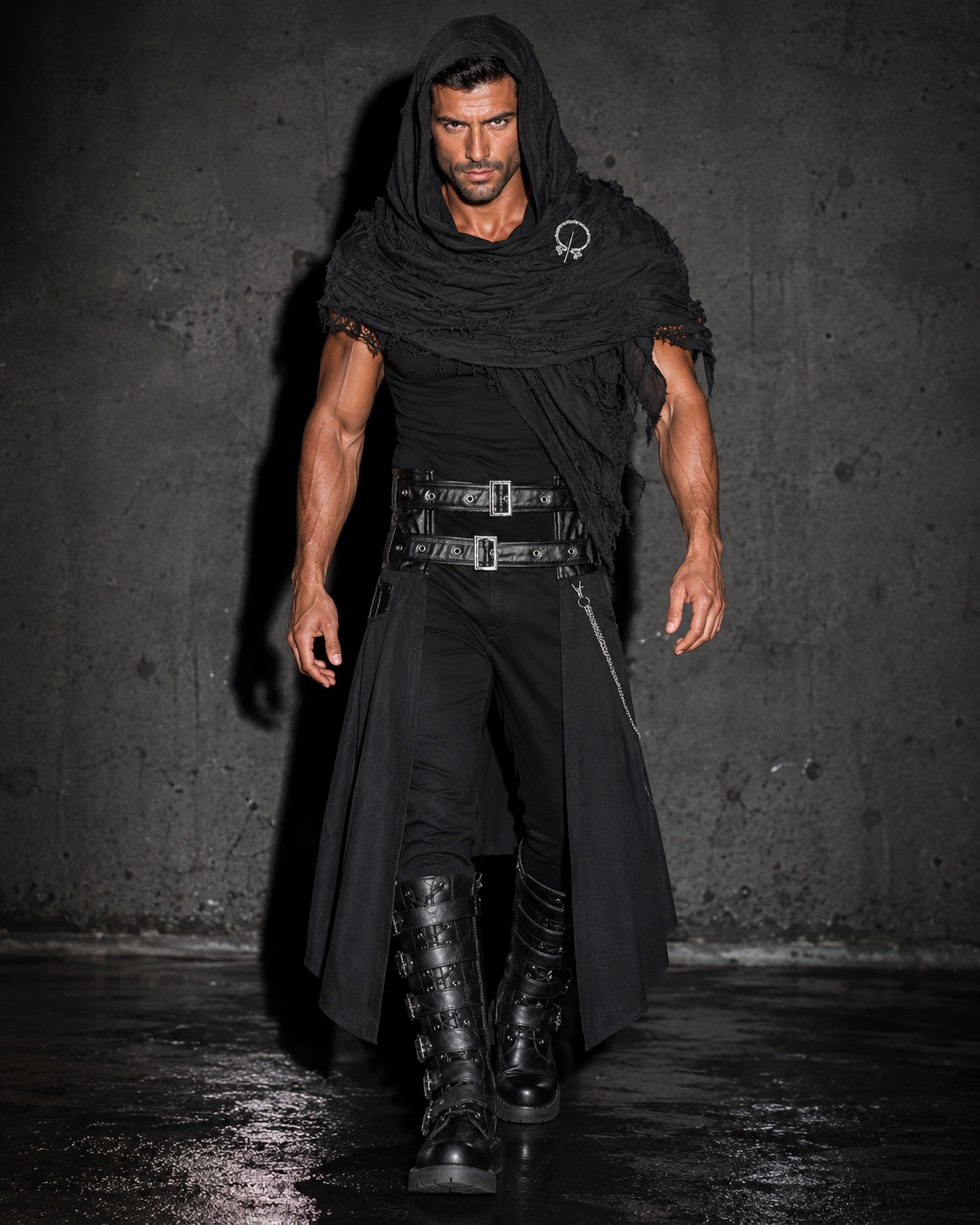 A man modeling a gothic black costume consisting of a hooded cape, a ruffled sleeveless overlay, a wide double-buckle belt, black pants, and tall lace-up boots, set against a dark textured concrete wall.