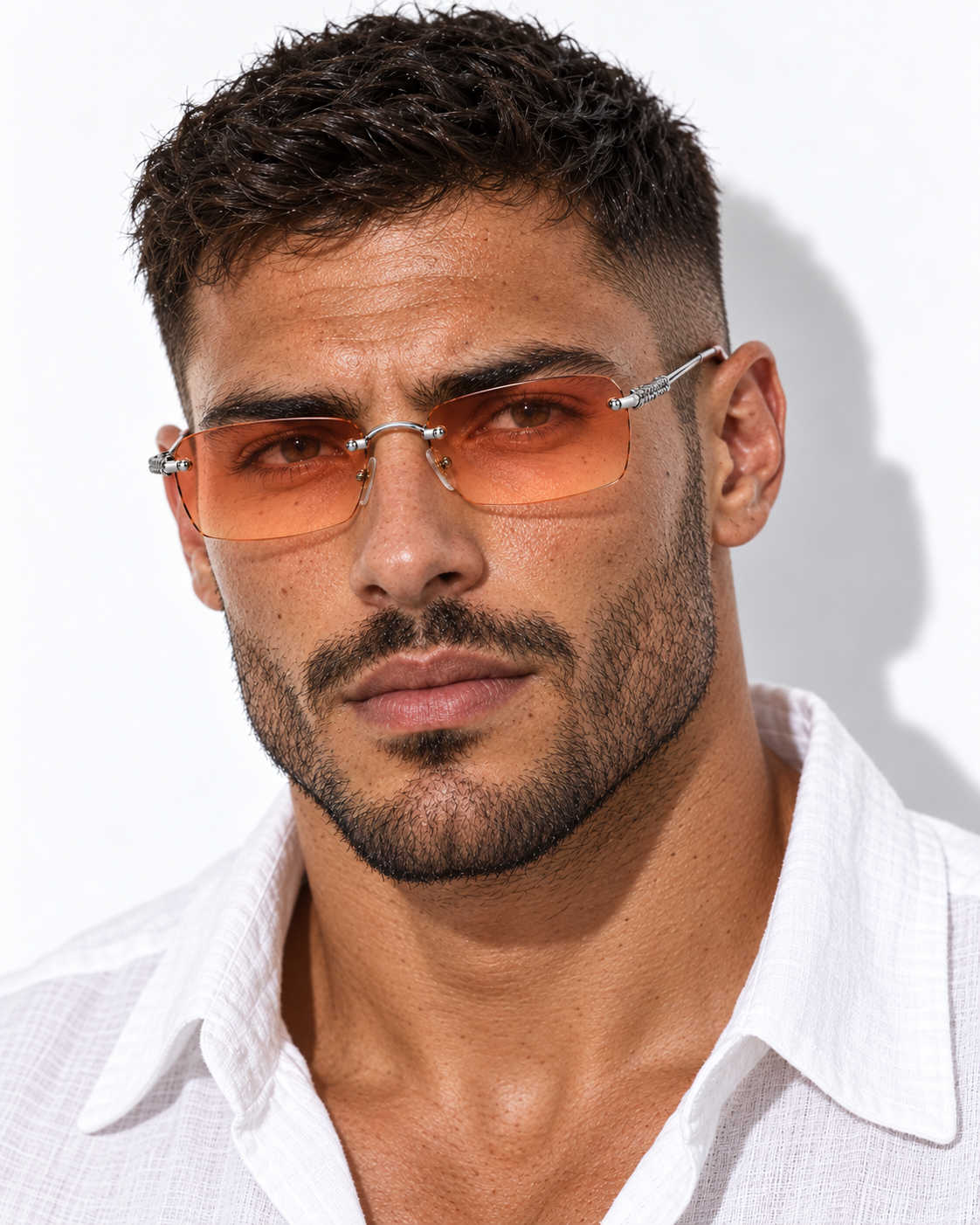 A close-up of a man wearing rimless square sunglasses with orange-tinted lenses and silver metal frames, paired with a white collared shirt against a white background.
