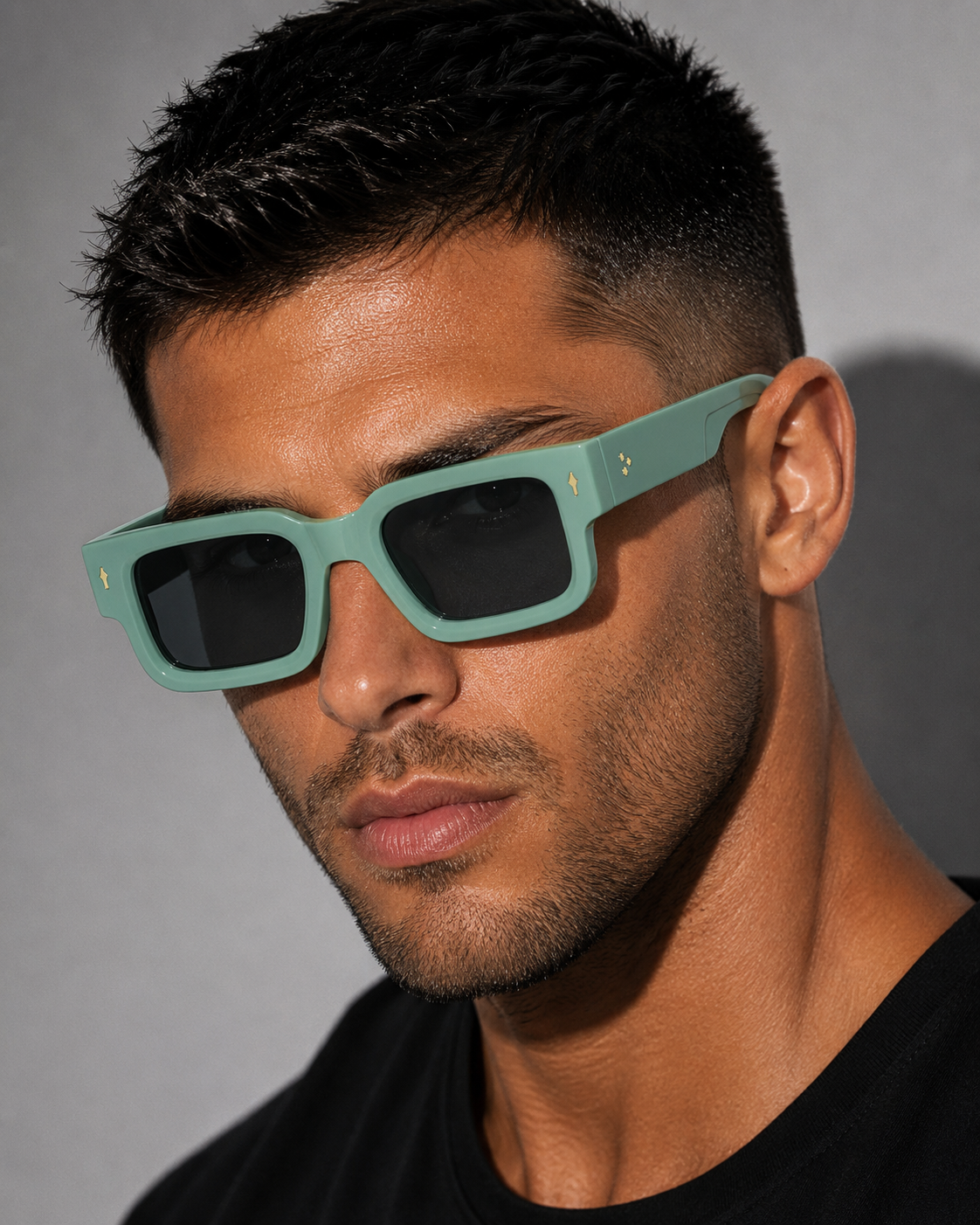 A close-up of a man wearing mint green square sunglasses with dark lenses and small gold logo accents on the temples, set against a neutral gray background.