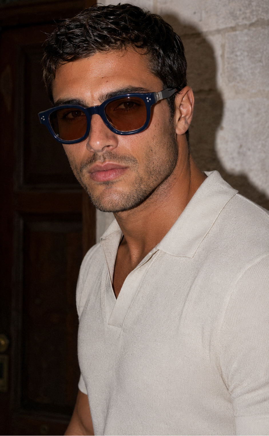 A man wearing thick blue-framed sunglasses with dark brown lenses and a white polo shirt, standing against a stone wall.