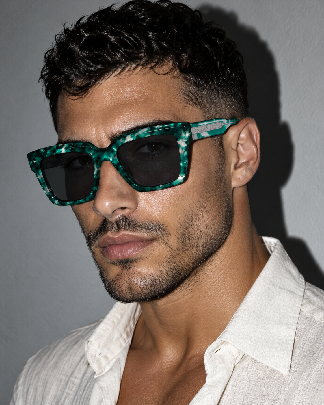 A close-up of a man wearing chunky square sunglasses with a green and white marbled pattern on the frames and dark lenses, paired with an open white shirt against a gray background.