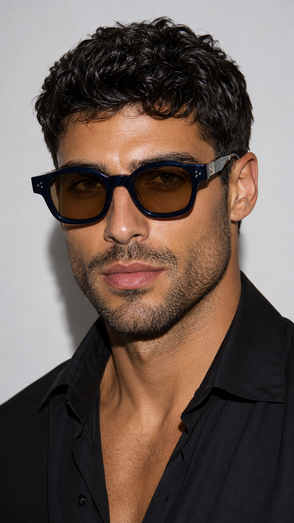 A close-up portrait of a man with dark curly hair wearing dark brown tinted sunglasses with a black frame and a black button-down shirt.